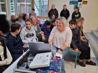 Frau Schieder sitzt am Laptop, im Hintergrund Schüler und Lehrpersonal